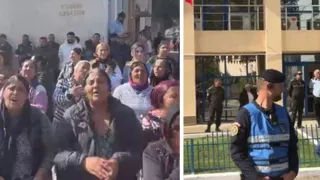 Un grup de persoane stă strâns într-un loc public, în timp ce un polițist în uniformă albastră și neagră stă în fața unei clădiri.