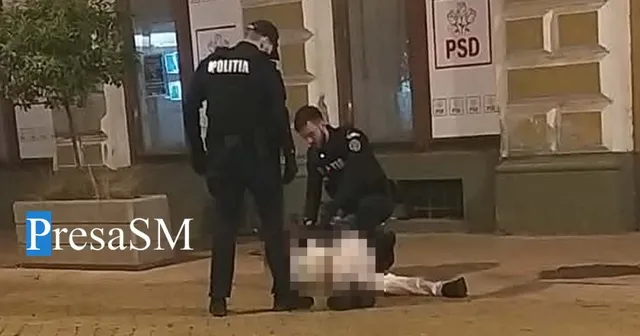 Un polițist în uniformă neagră stă în picioare lângă un alt polițist care imobilizează un bărbat întins pe trotuar în fața unei clădiri.