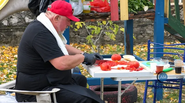 Un bărbat cu șapcă roșie și prosop alb pe gât taie roșii pe o masă albă lângă un pahar și un bol.