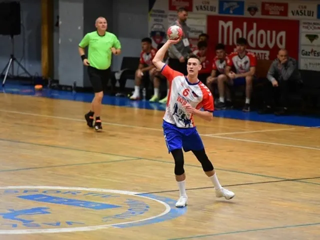 Un jucător de handbal în echipament albastru, roșu și alb ține o minge în mâna dreaptă pe un teren de sport cu spectatori și un arbitru în fundal.