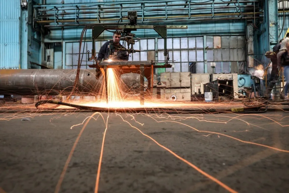 Un bărbat lucrează la o mașinărie industrială care scoate scântei strălucitoare deasupra unui cilindru metalic mare într-o fabrică.