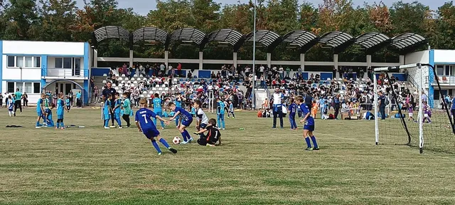 Câțiva jucători de fotbal în echipamente albastre joacă pe un teren de iarbă în fața unei tribune populate de spectatori.