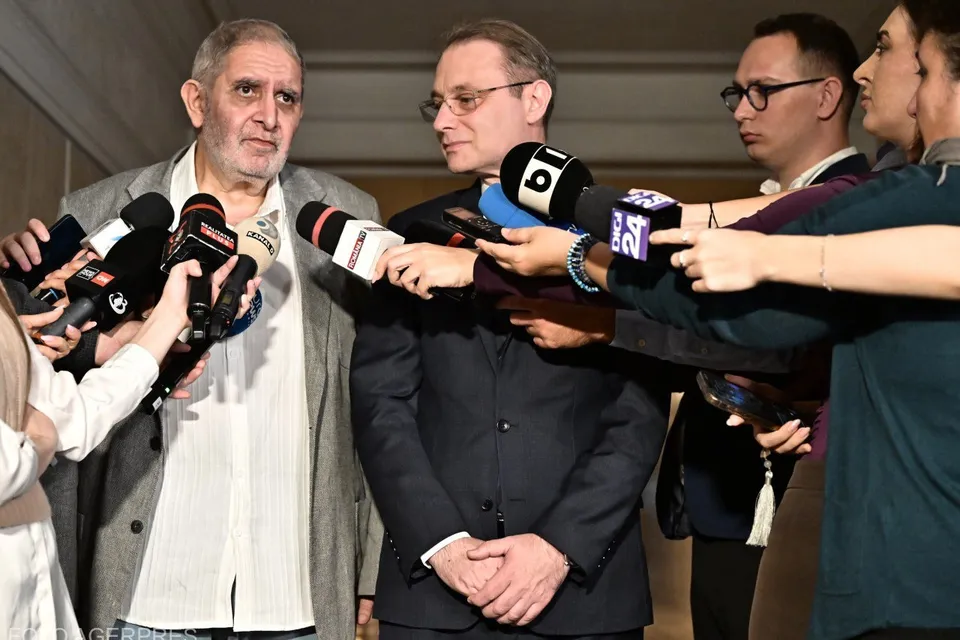 Un bărbat cu ochelari și sacou închis stă în fața mai multor microfoane ținute de persoane care îl înconjoară.