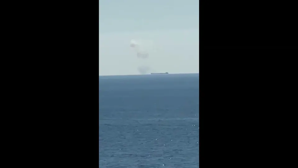 O navă de mare dimensiune este vizibilă la orizont pe o suprafață de apă sub un cer senin, emanând un nor de fum alb.