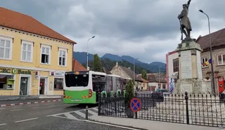 Un autobuz alb cu detalii verzi circulă pe un drum asfaltat lângă o clădire galbenă și un monument cu o statuie sub un cer noros.