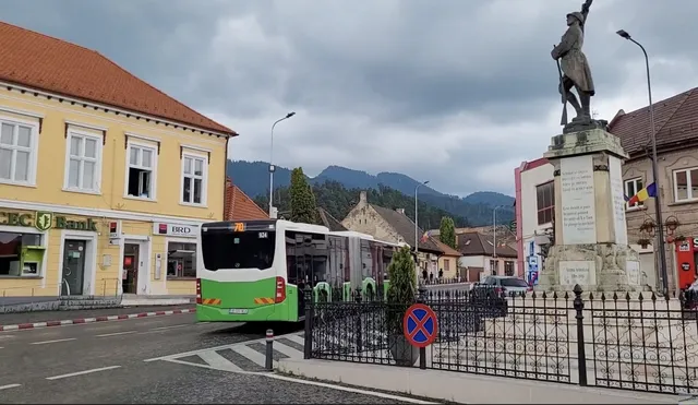 Un autobuz alb cu detalii verzi circulă pe un drum asfaltat lângă o clădire galbenă și un monument cu o statuie sub un cer noros.