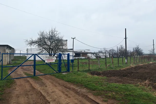 Un gard albastru de metal închide un drum de pământ lângă un câmp verde și câteva clădiri albe sub un cer înnorat.
