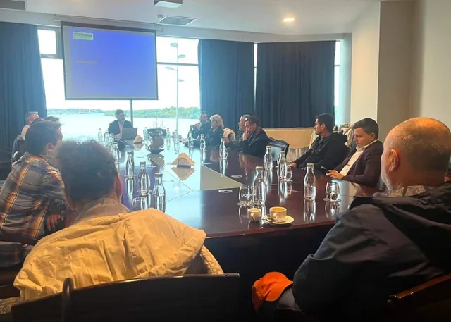 Oamenii stau la o masă lungă de conferință într-o încăpere cu un ecran de proiecție și ferestre mari în fundal.