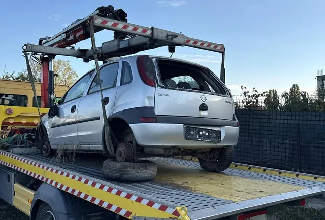 Un autoturism argintiu este ridicat de pe platforma unui camion cu ajutorul unei macarale metalice sub un cer senin.