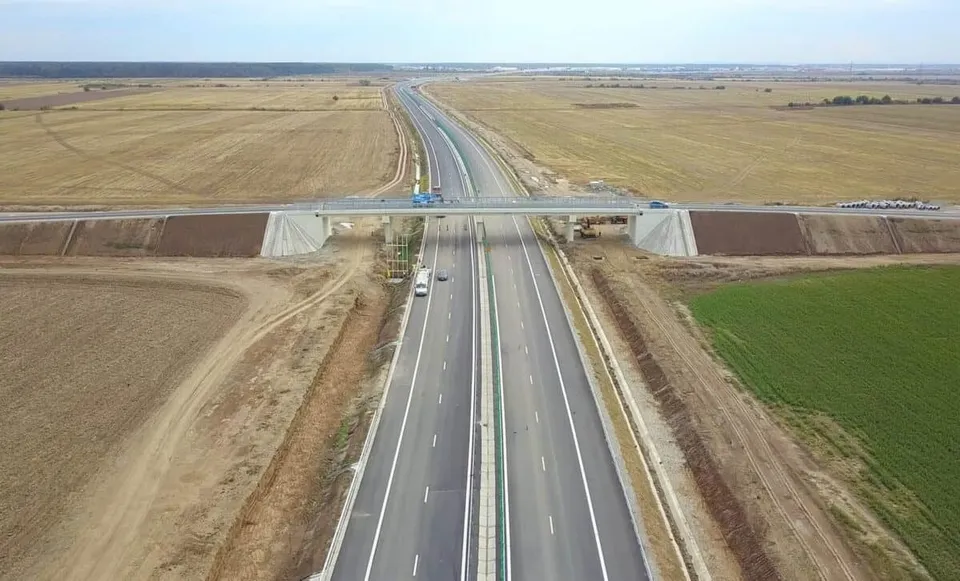O autostradă cu mai multe benzi de circulație traversează un câmp deschis, fiind traversată de un pod de o altă cale de acces.