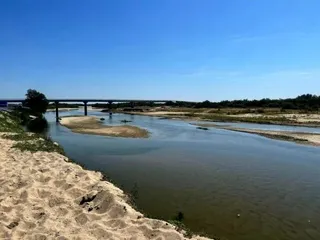 Un râu cu ape calme curge printre maluri nisipoase sub un cer senin, având un pod susținut de piloni în fundal.