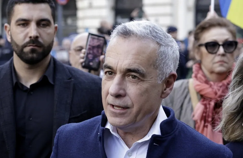 Un bărbat cu părul cărunt și sacou albastru este în centrul imaginii, fiind înconjurat de alte persoane în exterior.