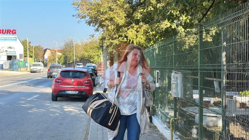 O femeie cu părul roșcat poartă un sac negru și un rucsac, mergând pe un trotuar lângă un gard de sârmă și mașini parcate.