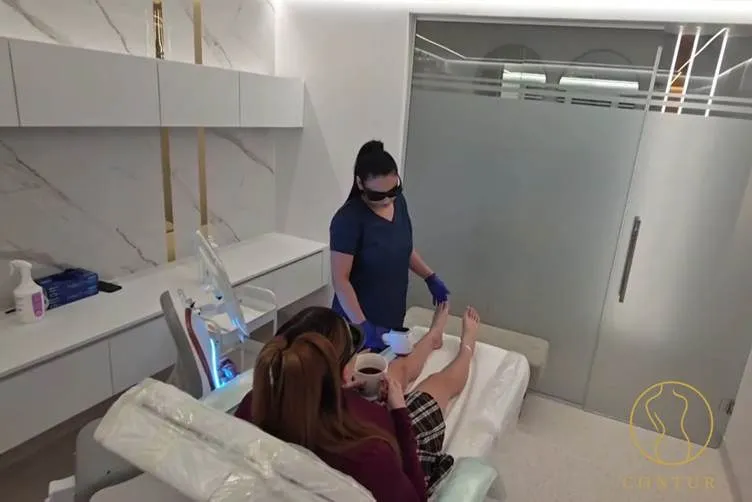O femeie cu haine închise la culoare și ochelari de protecție aplică un tratament pe piciorul unei persoane întinse pe o masă de tratament într-o cameră albă.