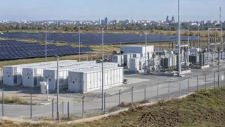 Rânduri de panouri fotovoltaice sunt instalate pe un câmp deschis, lângă containere albe de stocare și echipamente electrice protejate de un gard metalic.