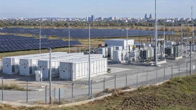 Rânduri de panouri fotovoltaice sunt instalate pe un câmp deschis, lângă containere albe de stocare și echipamente electrice protejate de un gard metalic.