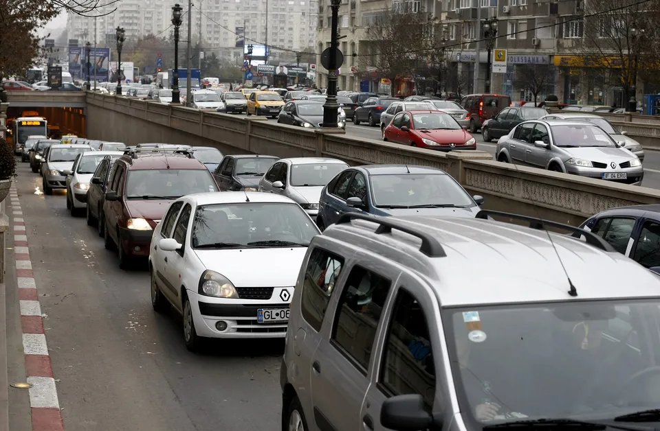 Mulți autovehicule sunt așezate în rânduri pe o stradă urbană lângă un gard metalic și clădiri înalte.