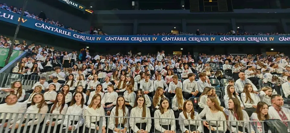 O mulțime de copii și tineri îmbrăcați în haine albe stau așezați pe scaune în tribunele unei arene închise.