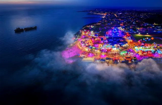 O vedere aeriană nocturnă a unei zone litorale iluminate cu lumini colorate, de lângă o mare întunecată unde plutesc două nave.