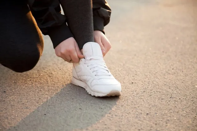 O persoană purtând pantaloni negri și o jachetă neagră își ajustează un pantof sport alb pe piciorul drept pe o suprafață asfaltată.