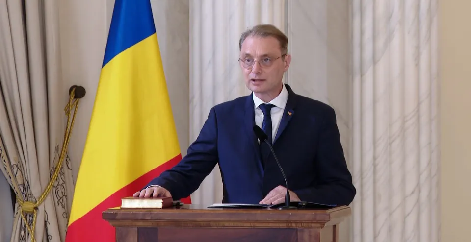 Un bărbat cu ochelari și costum închis la culoare stă în spatele unui podium de lemn lângă un steag tricolor.