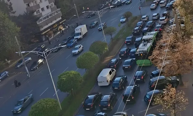 O vedere de sus a unui drum urban cu mai multe mașini circulând și parcate pe marginea unei benzi de vegetație cu copaci.