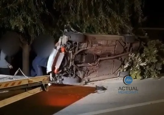 O mașină este răsturnată pe partea laterală lângă un arbust, cu partea inferioară expusă, în timp ce o platformă de tractare se află în prim plan.