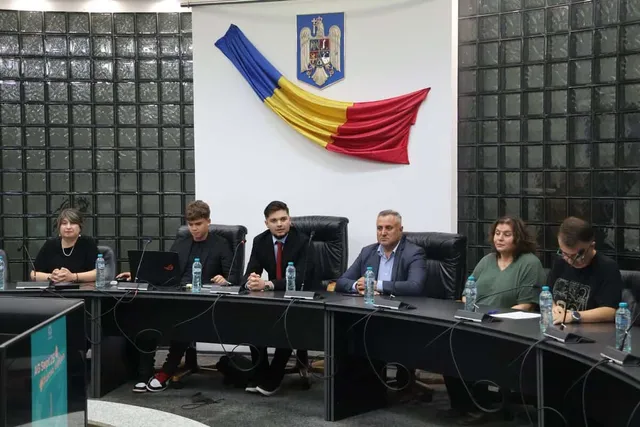 Șase persoane stau așezate la o masă curbată neagră în fața unui perete alb cu un steag și un blazon.