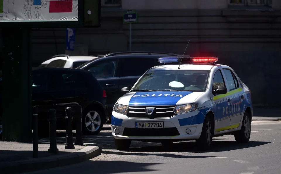O mașină de poliție albă și albastră cu girofarul aprins este parcată pe un drum lângă alte două autoturisme în fața unei clădiri.