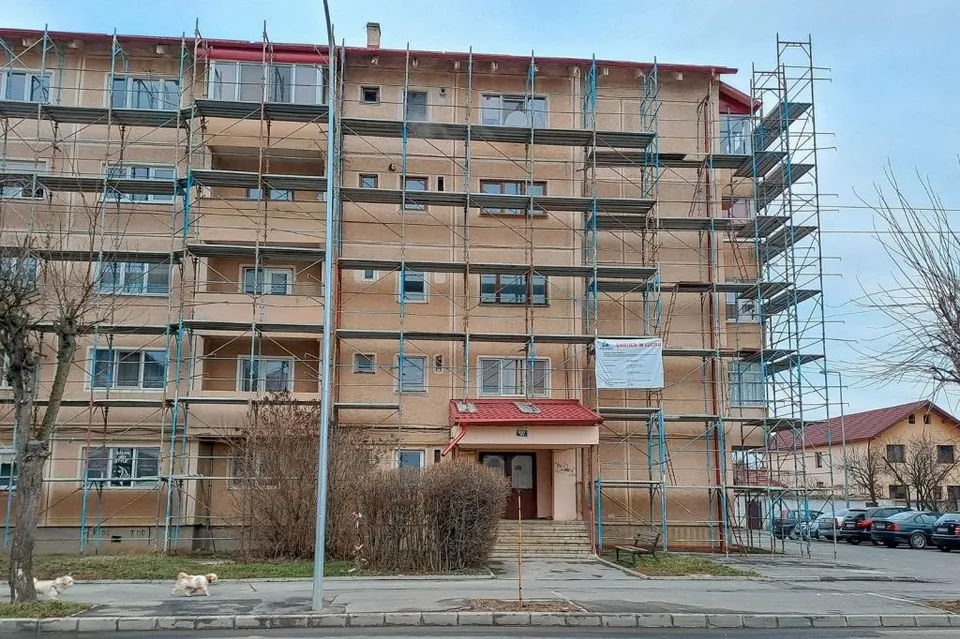 Un bloc de apartamente de culoare bej este acoperit de schele metalice pe mai multe niveluri sub un cer înnorat.