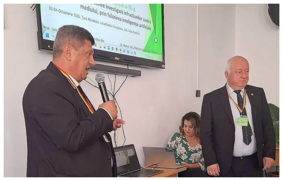 Un bărbat într-un costum negru vorbește într-un microfon în fața unui ecran de proiectare, în timp ce alți doi oameni stau în spatele unei mese.