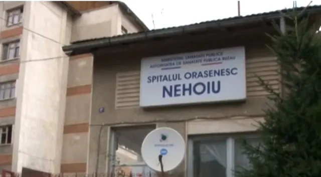 O signalație albă cu textul SPITALUL ORASENESC NEHOIU este montată pe fațada unei clădiri lângă un arbore conifer.