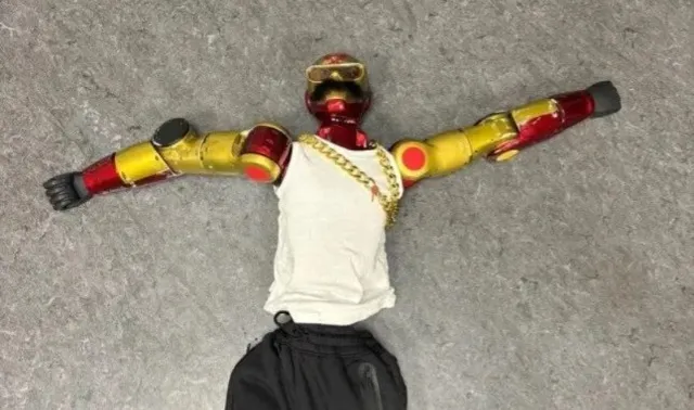 Un robot cu brațe de culoare roșie și galbenă poartă un tricou alb și un lanț auriu, fiind așezat pe un podea gri.