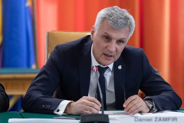 Un bărbat cu părul cărunt, purtând sacou închis la culoare și cravată, stă la o masă cu un microfon în față.
