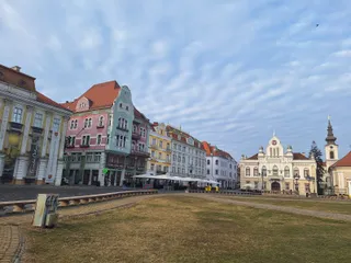 Clădiri colorate cu acoperișuri înclinate sunt dispuse pe o stradă lângă o zonă de prăpastie cu iarbă uscată sub un cer cu nori subțiri.