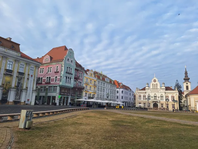 Clădiri colorate cu acoperișuri înclinate sunt dispuse pe o stradă lângă o zonă de prăpastie cu iarbă uscată sub un cer cu nori subțiri.