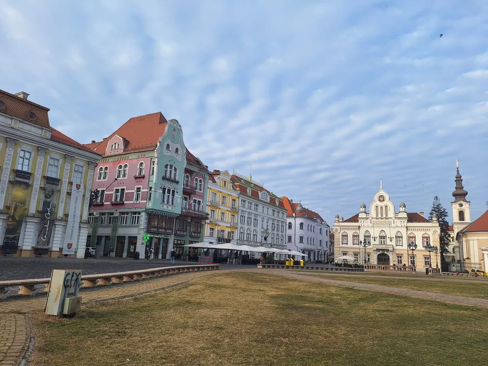 Clădiri colorate cu acoperișuri înclinate sunt dispuse pe o stradă lângă o zonă de prăpastie cu iarbă uscată sub un cer cu nori subțiri.