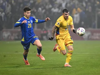 Doi jucători de fotbal, unul în echipament albastru și unul în echipament galben, aleargă pe un teren de fotbal lângă un balon.