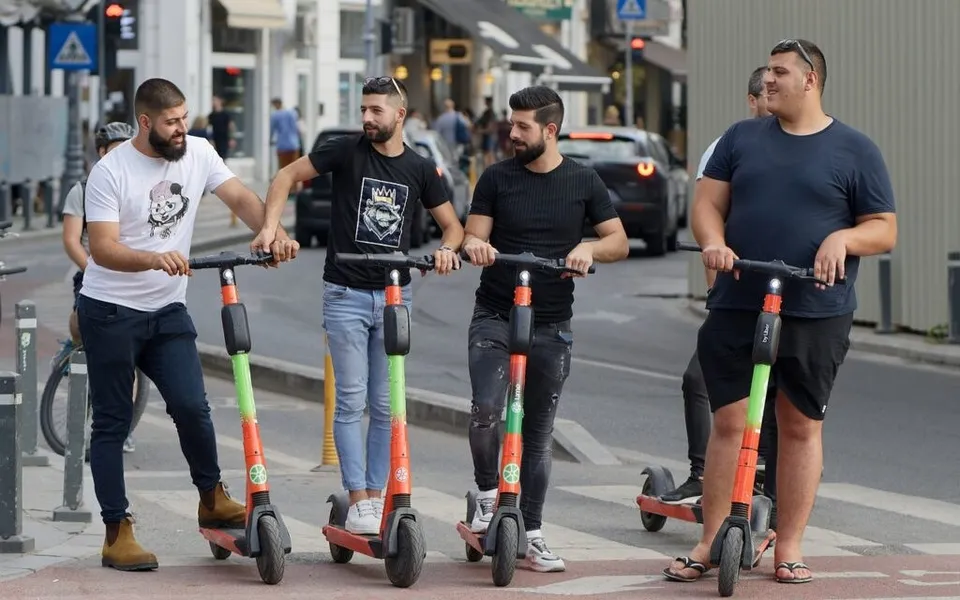 Patru bărbați stau pe trotinete electrice colorate pe un trotuar lângă o stradă cu mașini și clădiri.