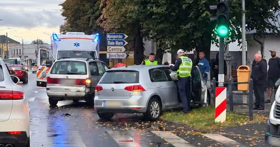 Patru autoturisme și o ambulanță sunt oprite pe un drum umed lângă un semafor cu lumină verde și un trotuar cu un coș de gunoi galben.