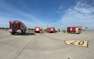 Mai multe mașini de pompieri roșii sunt parcate pe o pistă de beton sub un cer albastru cu nori subțiri.