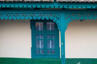 O fereastră cu rame verzi este situată sub un acoperiș decorat cu elemente de lemn sculptat de aceeași culoare pe un perete deschis la culoare.