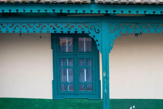 O fereastră cu rame verzi este situată sub un acoperiș decorat cu elemente de lemn sculptat de aceeași culoare pe un perete deschis la culoare.