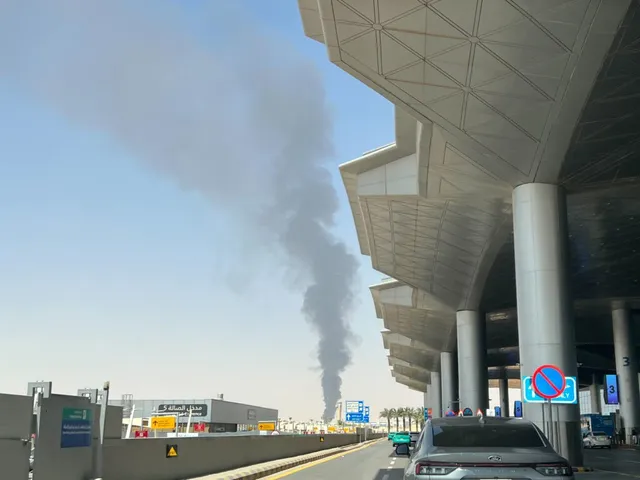 Un fir de fum negru se ridică vertical spre cer de lângă un terminal de aeroport cu acoperiș metalic și coloane cilindrice gri.