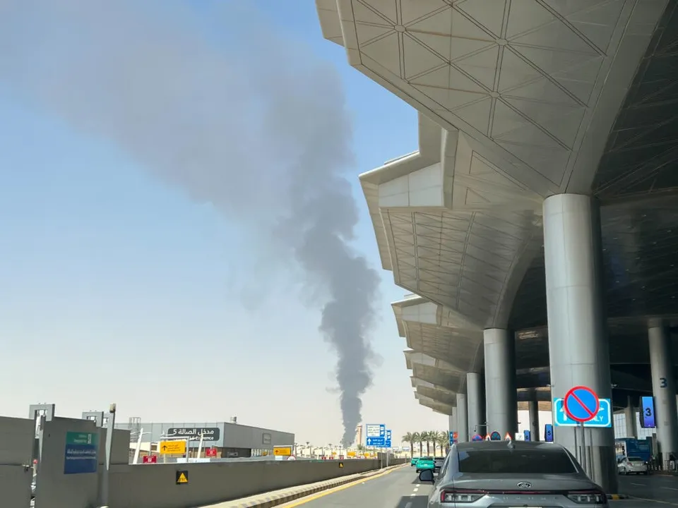 Un fir de fum negru se ridică vertical spre cer de lângă un terminal de aeroport cu acoperiș metalic și coloane cilindrice gri.
