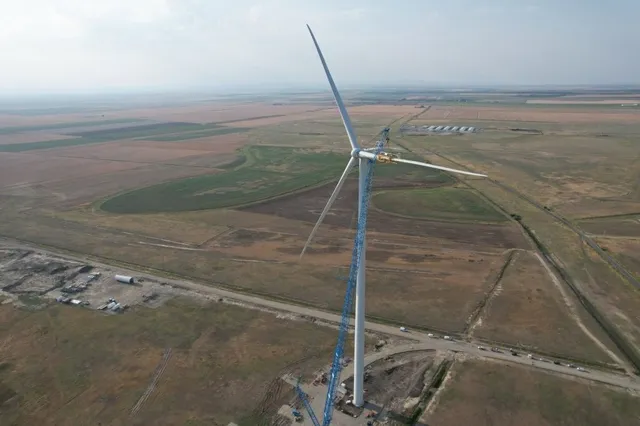 O turbină eoliană albă este situată pe un câmp deschis lângă un drum de țară, sub un cer înnorat.