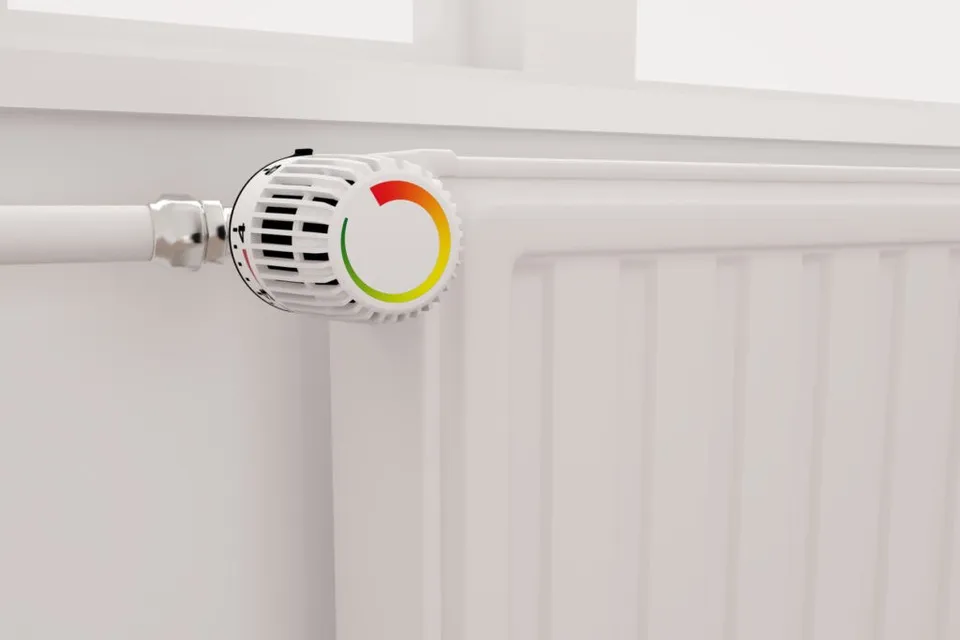Un radiator alb este conectat la o conductă prin intermediul unui termostat cu un indicator colorat în roșu, galben și verde.