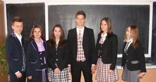 Șase tineri stau în rând în fața unei table negre, purtând sacouri și fuste sau pantaloni cu modele în carouri sau culori unies.