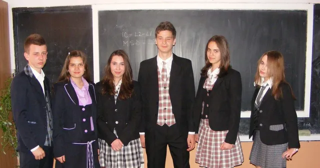 Șase tineri stau în rând în fața unei table negre, purtând sacouri și fuste sau pantaloni cu modele în carouri sau culori unies.