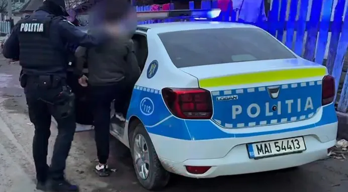 Un polițist în uniformă neagră stă lângă un vehicul alb cu inscripții albastre și un tânăr îmbrăcat în haine închise lângă mașină.